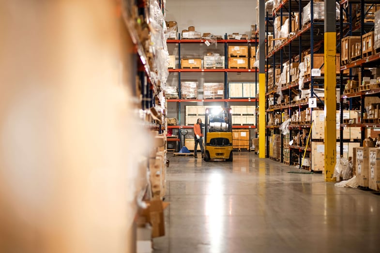Warehouse worker standing beside a forklift in a large industrial storage aisle with tall shelves filled with boxes and crates.