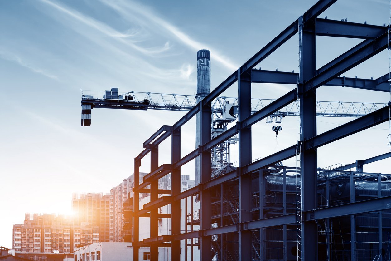 Steel framework of an industrial building under construction with cranes in the background, set against a city skyline and a bright sunrise.