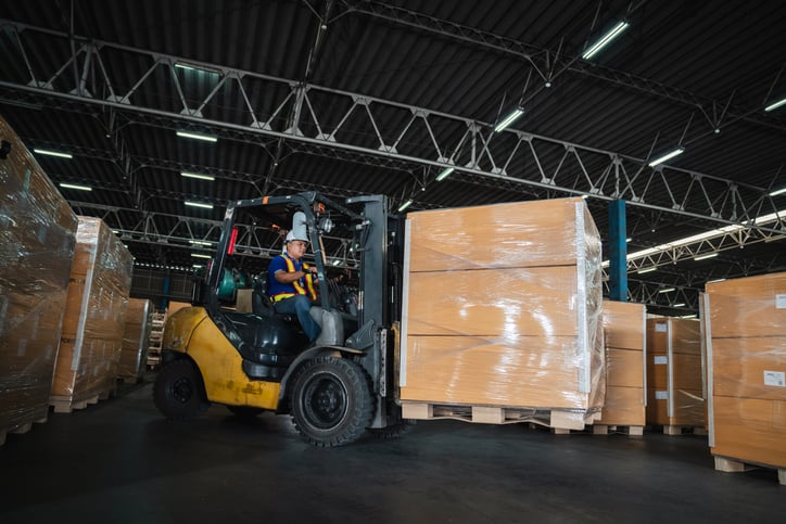 Forklift operator moving large palletized boxes inside a warehouse with organized inventory shelves and industrial lighting.