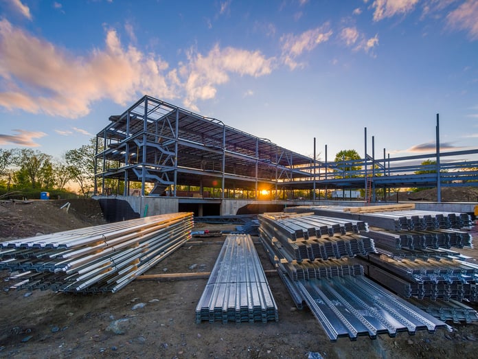 Wide-angle view of a steel-frame building under construction at sunset, with stacks of metal decking and construction materials in the foreground.