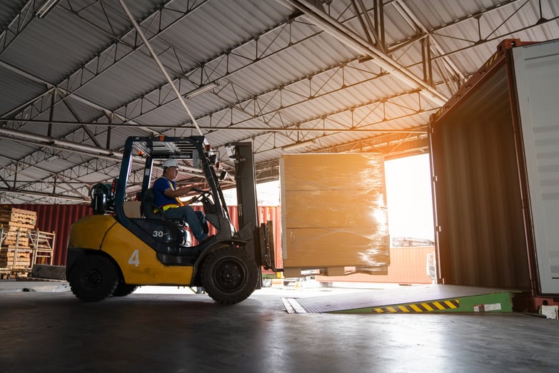 Forklift loading palletized construction materials into a shipping container inside an industrial warehouse, representing reliable logistics, direct-to-jobsite delivery, and efficient supply chain management for large-scale infrastructure and data center projects.
