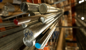 Threaded metal rods with red, blue, and dark-colored ends, arranged diagonally on a storage rack in an industrial setting.