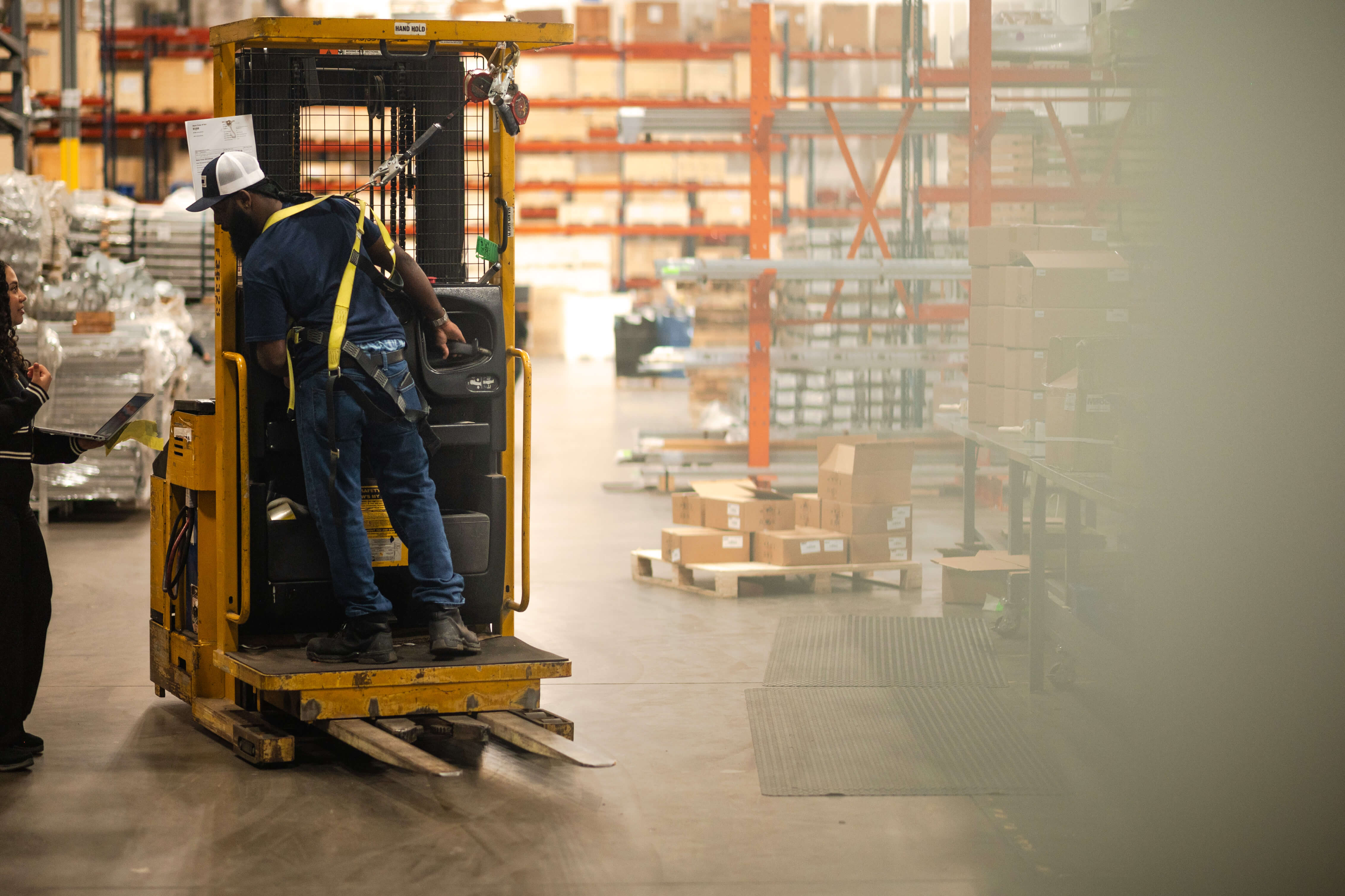 A warehouse worker in a safety harness operates a yellow lift truck while speaking with a colleague holding a laptop. They are surrounded by boxes, shelving, and industrial inventory inside a well-lit distribution center