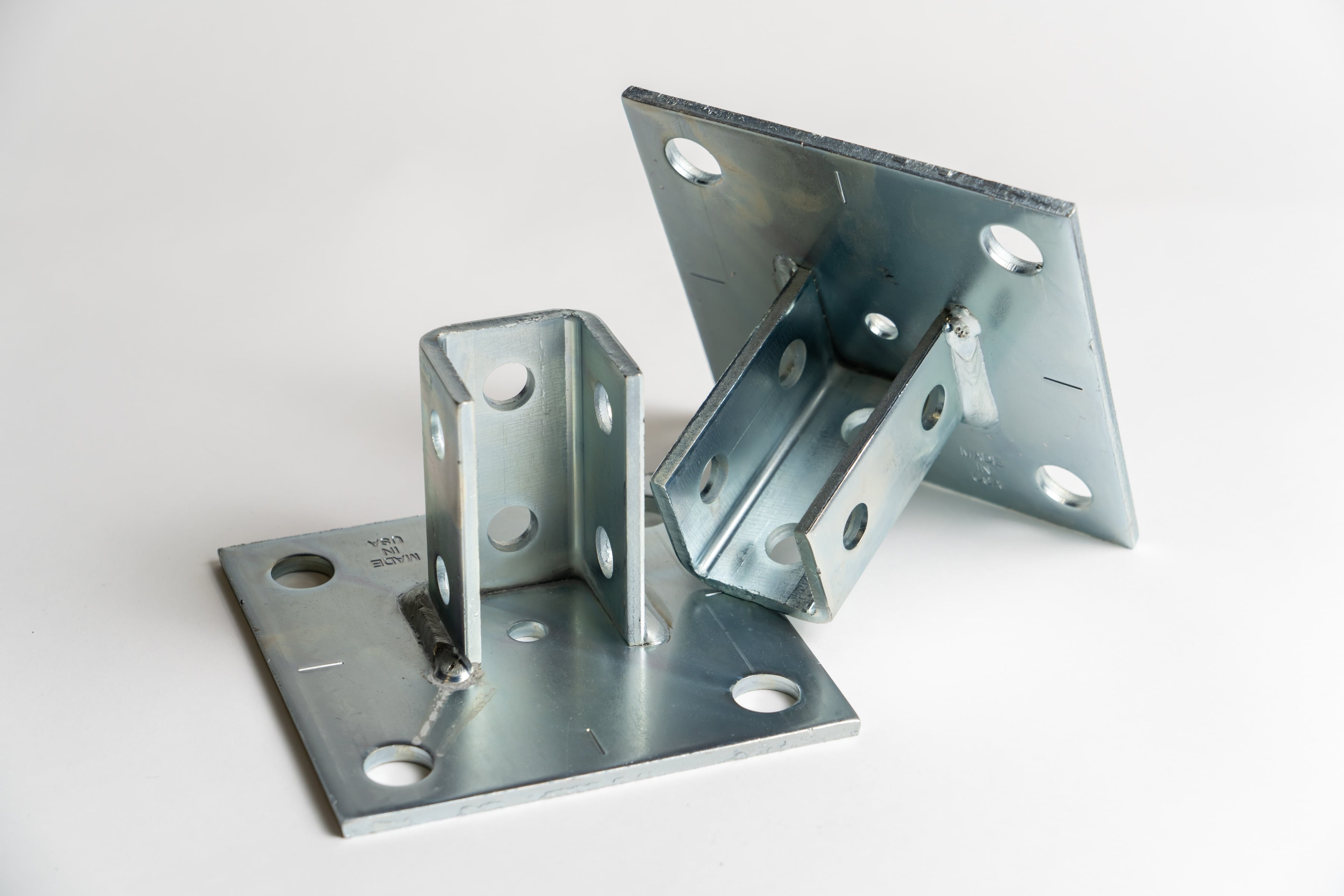 Two galvanized steel post bases for structural electrical applications, featuring square mounting plates with multiple anchor holes and welded U-shaped brackets for utility and infrastructure support.