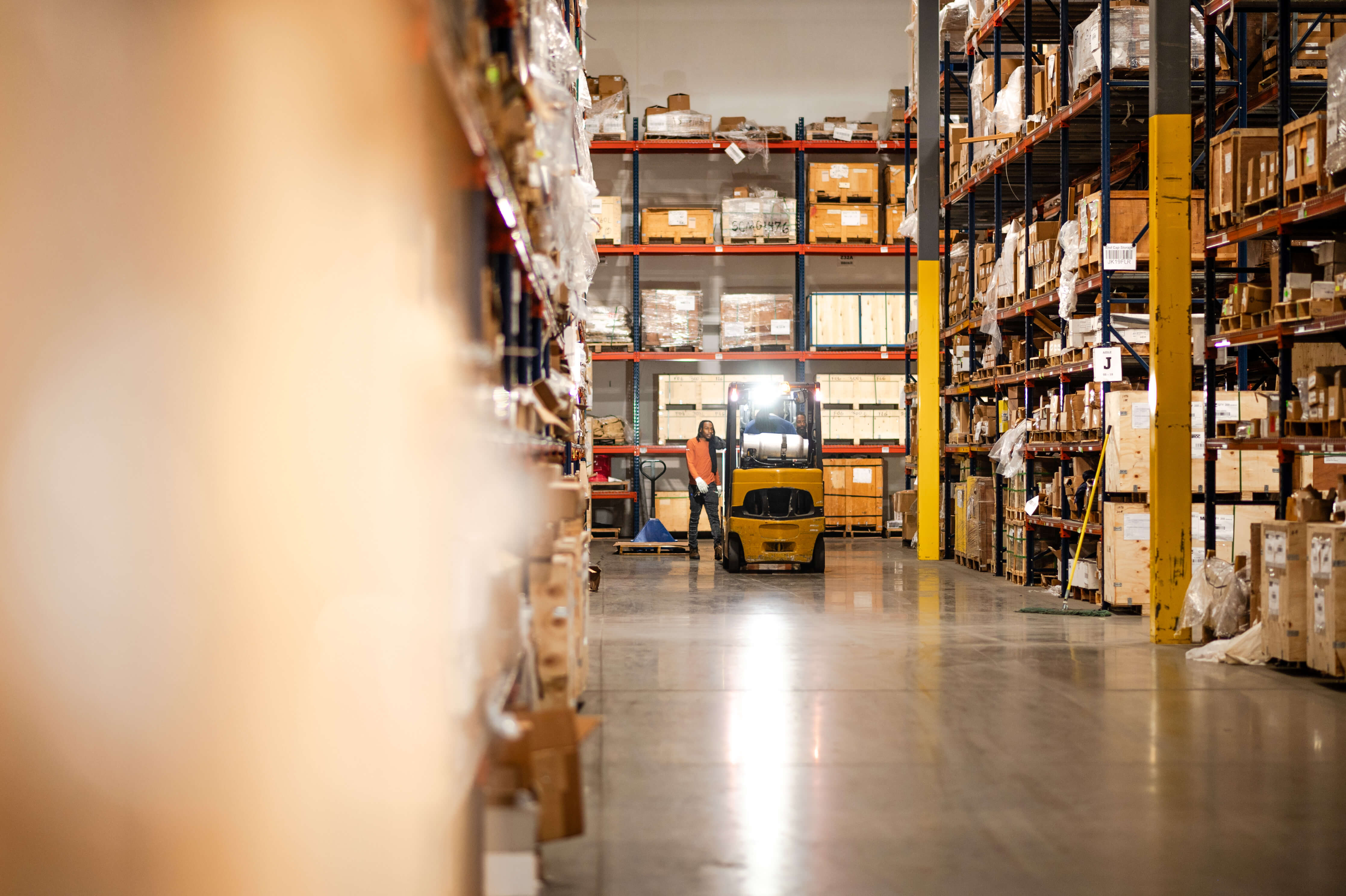 Warehouse worker standing beside a forklift in a large industrial storage aisle with tall shelves filled with boxes and crates.