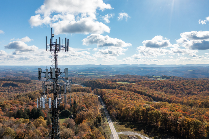 A tall telecommunications tower equipped with multiple antennas rises above a colorful autumn forest. A winding road cuts through the trees, and rolling hills stretch into the distance beneath a partly cloudy sky with bright sunlight.